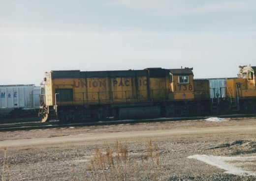 A yellow Union Pacific diesel locomotive numbered 1738 sits on tracks in a yard with snow patches on the ground, with a white freight car visible behind it.