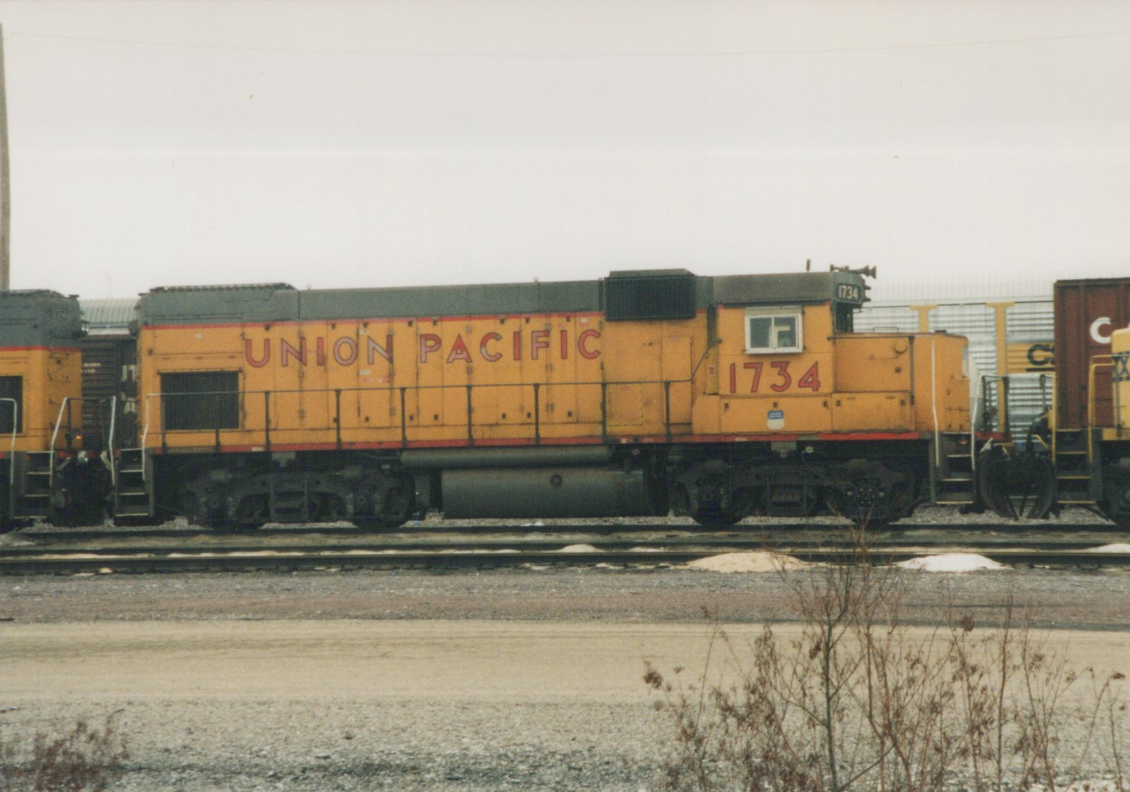 Side view of a yellow Union Pacific diesel locomotive number 1734, parked on tracks in a snowy yard with other railcars nearby.