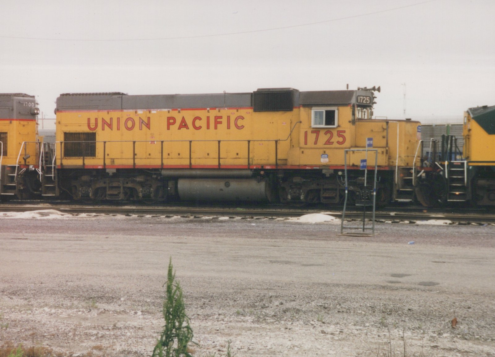Side view of a yellow Union Pacific diesel locomotive numbered 1725 in a snowy railroad yard, with another locomotive numbered 1700 partially visible behind it.