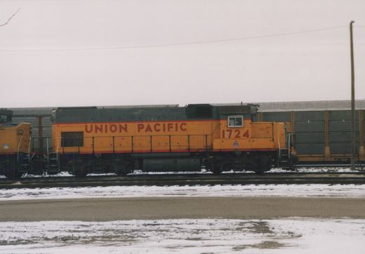 A yellow and grey Union Pacific diesel locomotive numbered 1724 stands on tracks in a snow-covered yard, with another locomotive partially visible behind it.