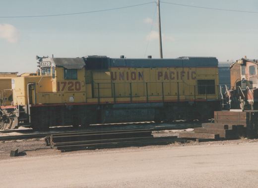 A yellow Union Pacific locomotive number 1720 sits on tracks in a rail yard with another locomotive partially visible in the background.