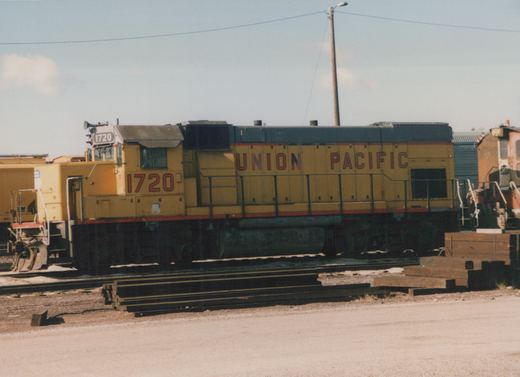 A yellow Union Pacific diesel locomotive numbered 1720 stands in a rail yard on a clear, sunny day with stacks of steel rails in the foreground.