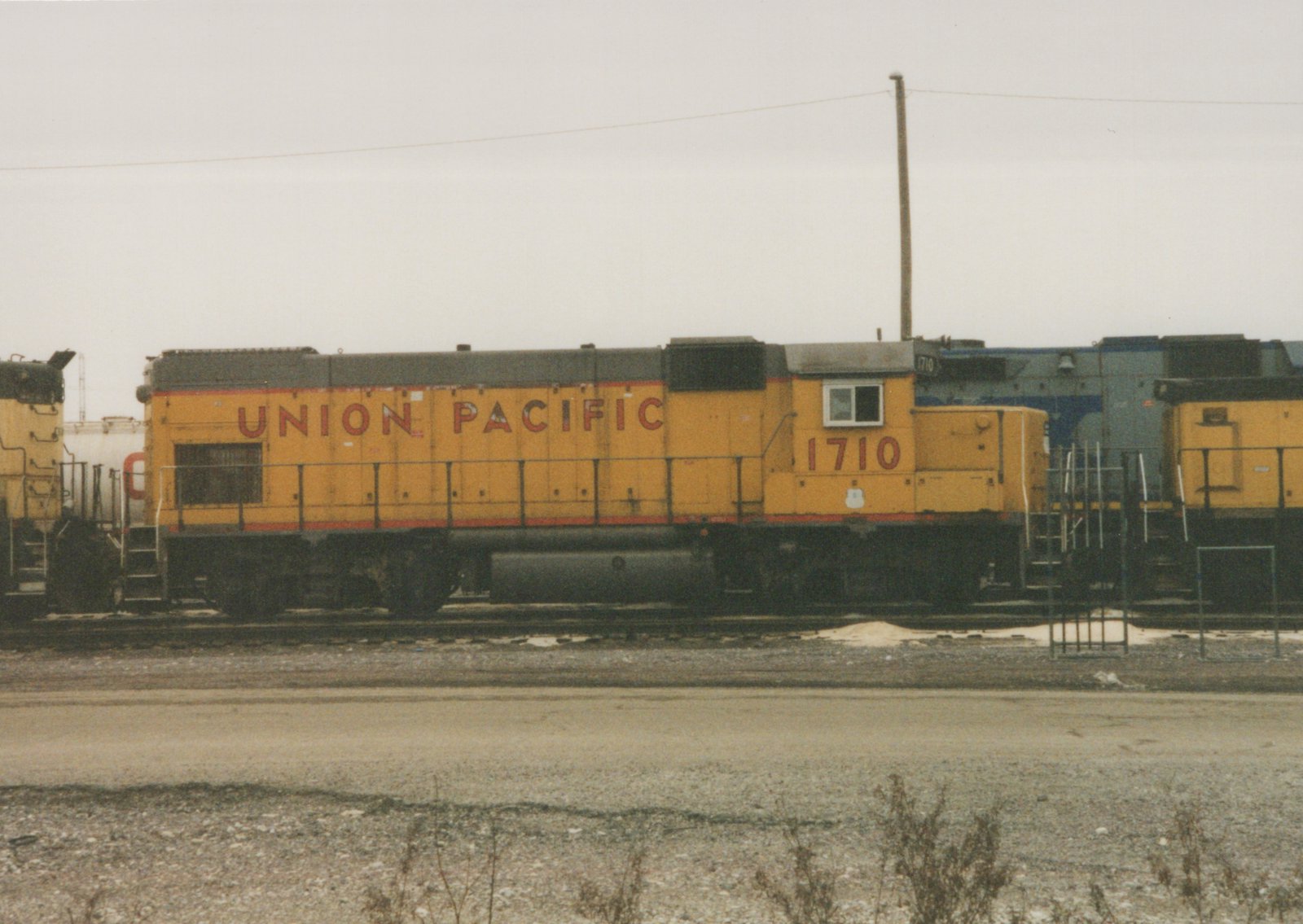 A side view of a yellow Union Pacific diesel locomotive, number 1710, standing in a rail yard. A blue locomotive is partially visible behind it, and a gravel foreground is in the bottom of the frame.