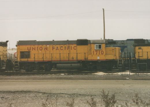 A side view of a yellow Union Pacific diesel locomotive, number 1710, standing in a rail yard. A blue locomotive is partially visible behind it, and a gravel foreground is in the bottom of the frame.