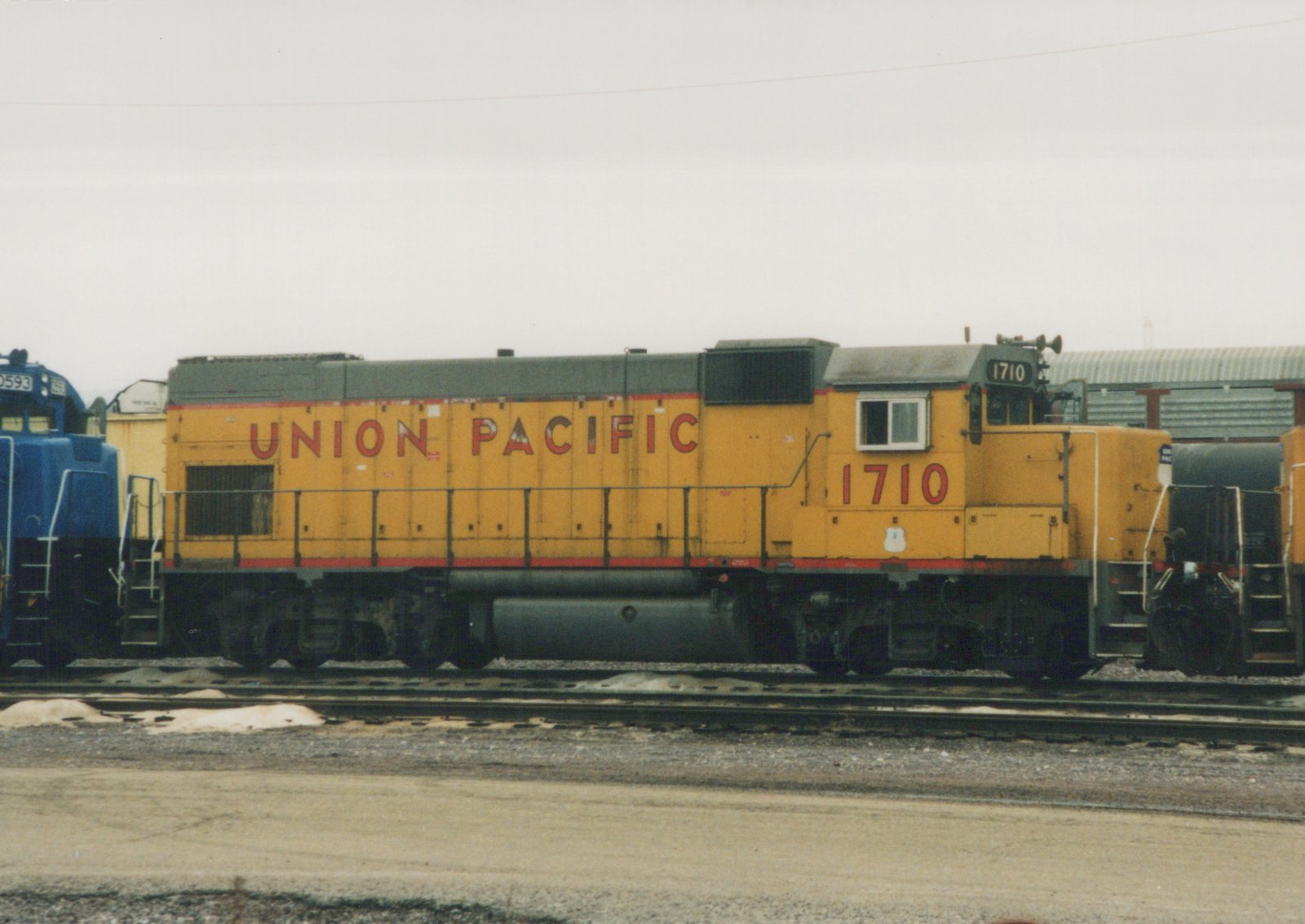 A yellow and gray Union Pacific diesel locomotive numbered 1710 is parked on tracks in a rail yard, with a blue locomotive and a black tank car nearby.