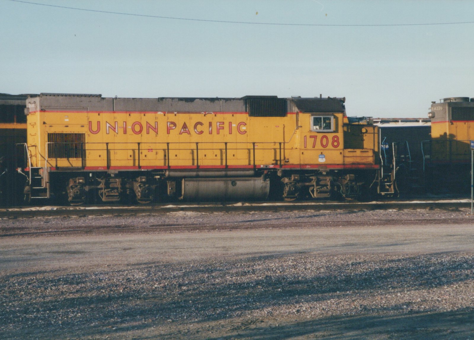 A side view of a yellow Union Pacific diesel locomotive, number 1708, parked on tracks at a rail yard on a clear day. A second locomotive is partially visible in the background.