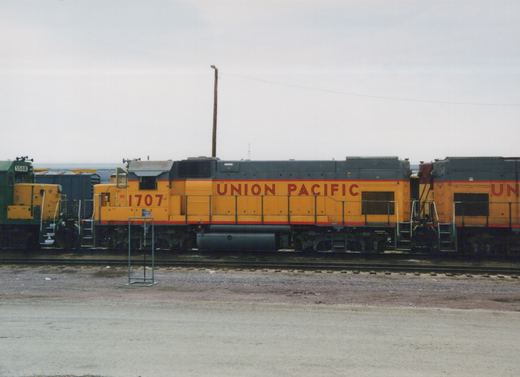 Side view of a yellow and gray Union Pacific diesel locomotive, number 1707, with other locomotives visible in the background, all sitting on tracks in a gravel yard under an overcast sky.