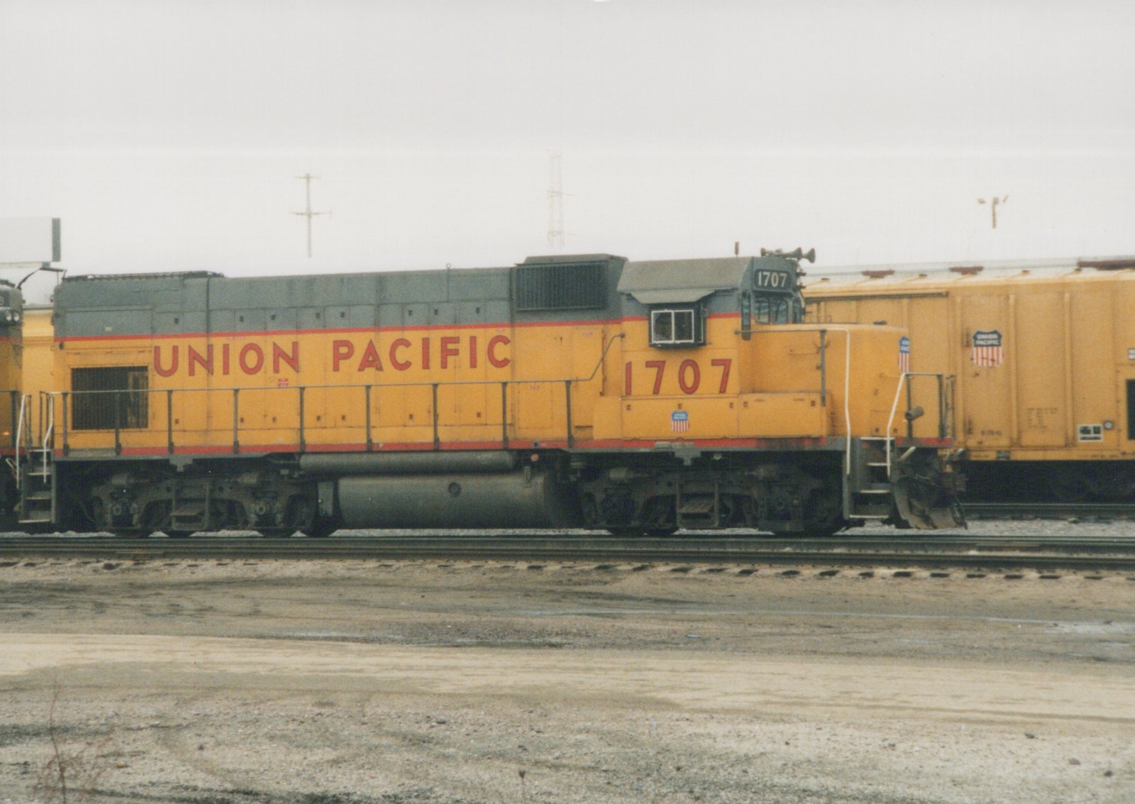 A side view of a yellow Union Pacific diesel locomotive number 1707 with a gray hood, parked on tracks in a gravel rail yard with an overcast sky.
