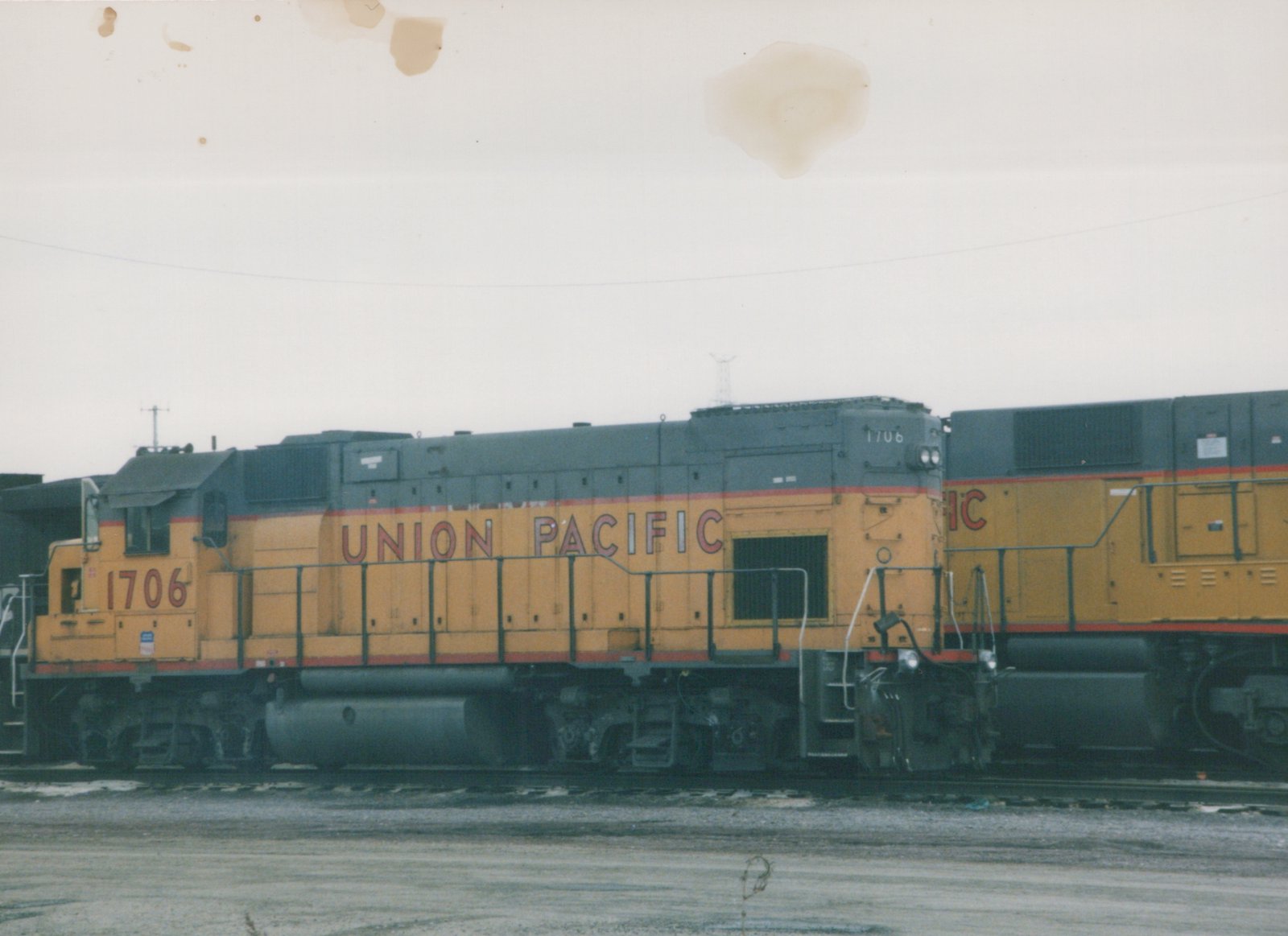 Side view of a Union Pacific diesel locomotive number 1706, painted yellow with a gray roof and red stripes, coupled to another locomotive in a rail yard on a gray, overcast day.