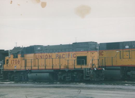 Side view of a Union Pacific diesel locomotive number 1706, painted yellow with a gray roof and red stripes, coupled to another locomotive in a rail yard on a gray, overcast day.