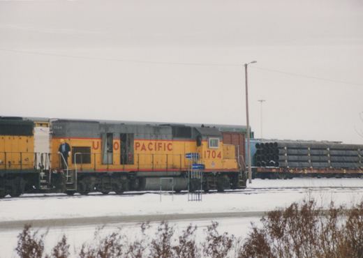 A yellow and gray Union Pacific locomotive numbered 1704 is parked on snowy tracks, with one person standing on its steps and freight cars loaded with pipes in the background.