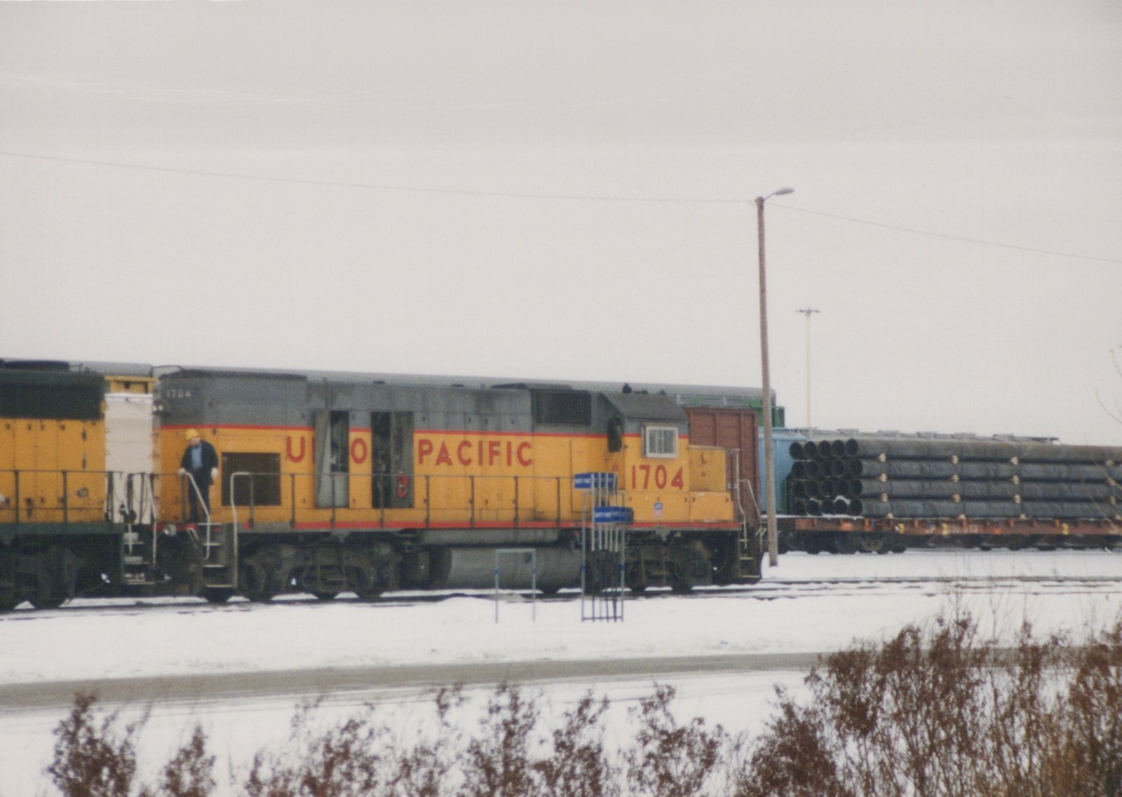 A yellow and gray Union Pacific locomotive numbered 1704 is parked on snowy tracks, with one person standing on its steps and freight cars loaded with pipes in the background.
