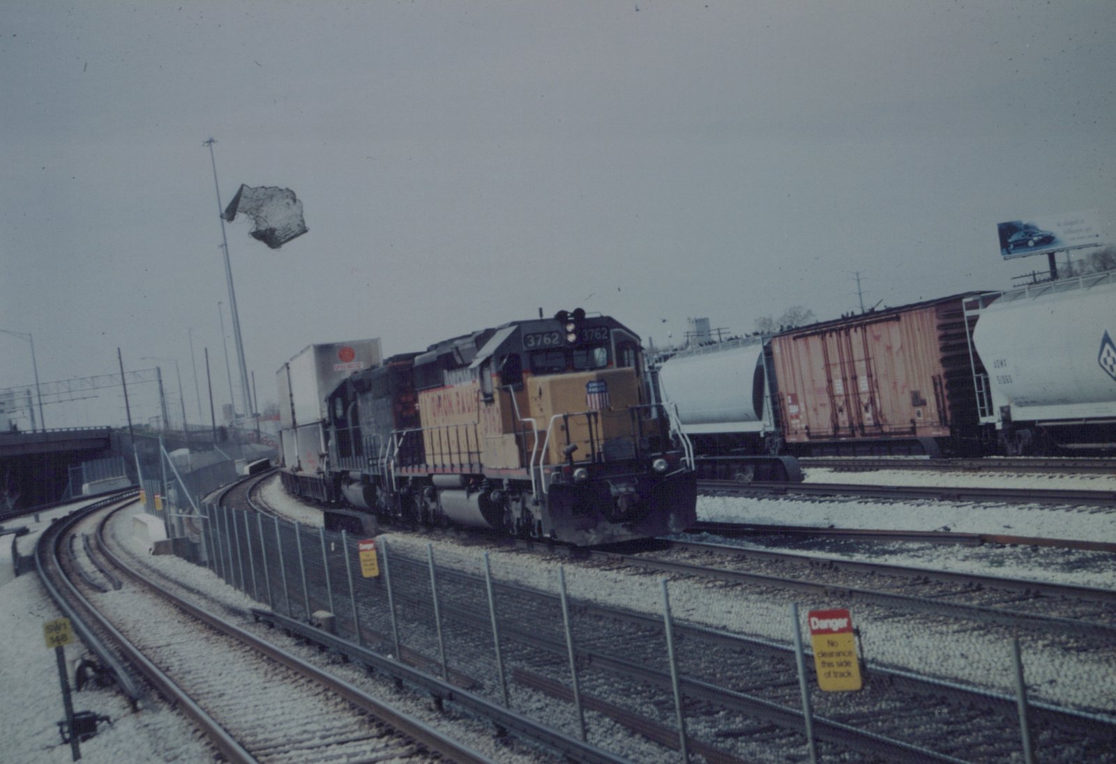 A yellow and gray Union Pacific diesel locomotive numbered 3762 is seen on the tracks with freight cars, surrounded by snow and a cloudy sky.