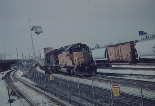 A yellow and gray Union Pacific diesel locomotive numbered 3762 is seen on the tracks with freight cars, surrounded by snow and a cloudy sky.