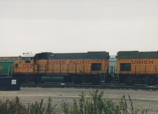 Two Union Pacific locomotives, with number 1702 clearly visible, sit on tracks in a rail yard with overgrown weeds in the foreground and a cloudy sky above.