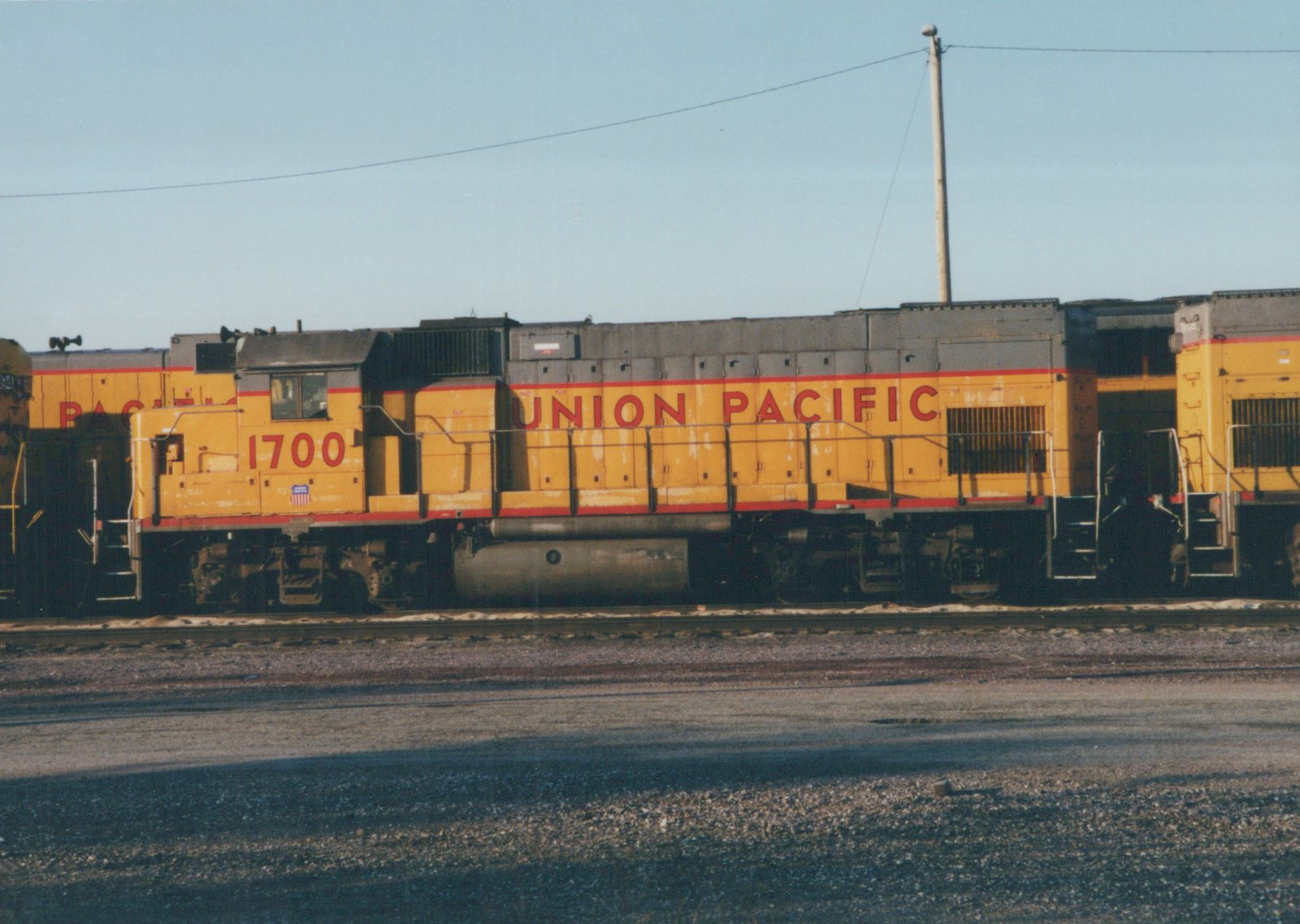 A yellow and red Union Pacific diesel locomotive numbered 1700 is parked on tracks in a gravel yard with a clear blue sky overhead.