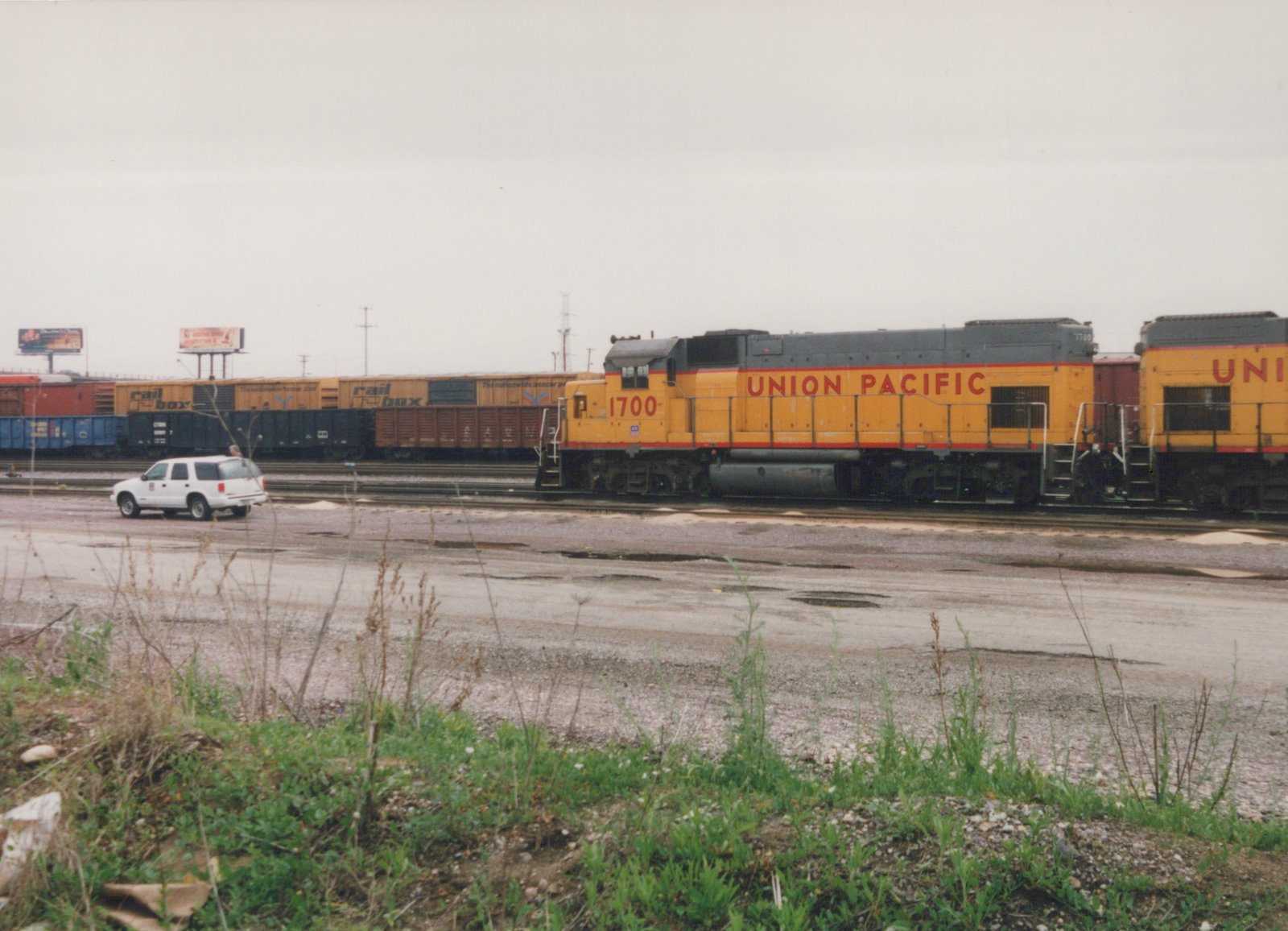 A side view of a yellow Union Pacific diesel locomotive, number 1700, pulling freight cars in a rail yard, with a white SUV parked nearby and weeds in the foreground.