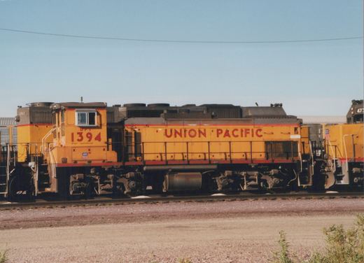 A yellow Union Pacific diesel locomotive numbered 1394 stands on tracks in a gravel yard, with other locomotives visible behind and beside it.