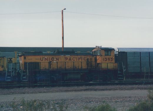 A yellow Union Pacific locomotive numbered 1393 is shown in side view on railroad tracks in a yard, with a dark freight car behind it and a building in the background.