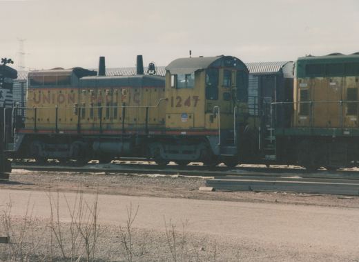 A yellow and gray Union Pacific diesel locomotive, number 1247, is sitting on tracks in a yard, with a second locomotive coupled behind it.
