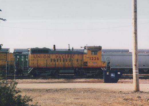 A yellow and black Union Pacific locomotive numbered 1226 stands on a track in a rail yard, with a silver tanker car and a green caboose in the background under an overcast sky.
