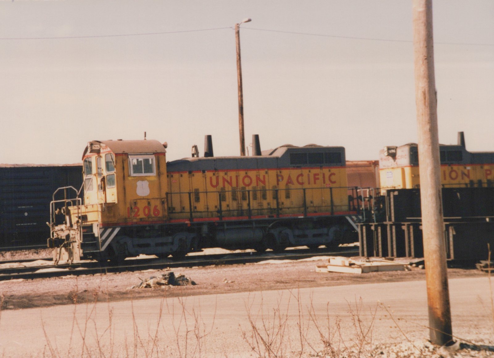 A yellow and gray Union Pacific diesel locomotive numbered 1206 is parked on tracks in a rail yard with another locomotive and freight cars in the background.