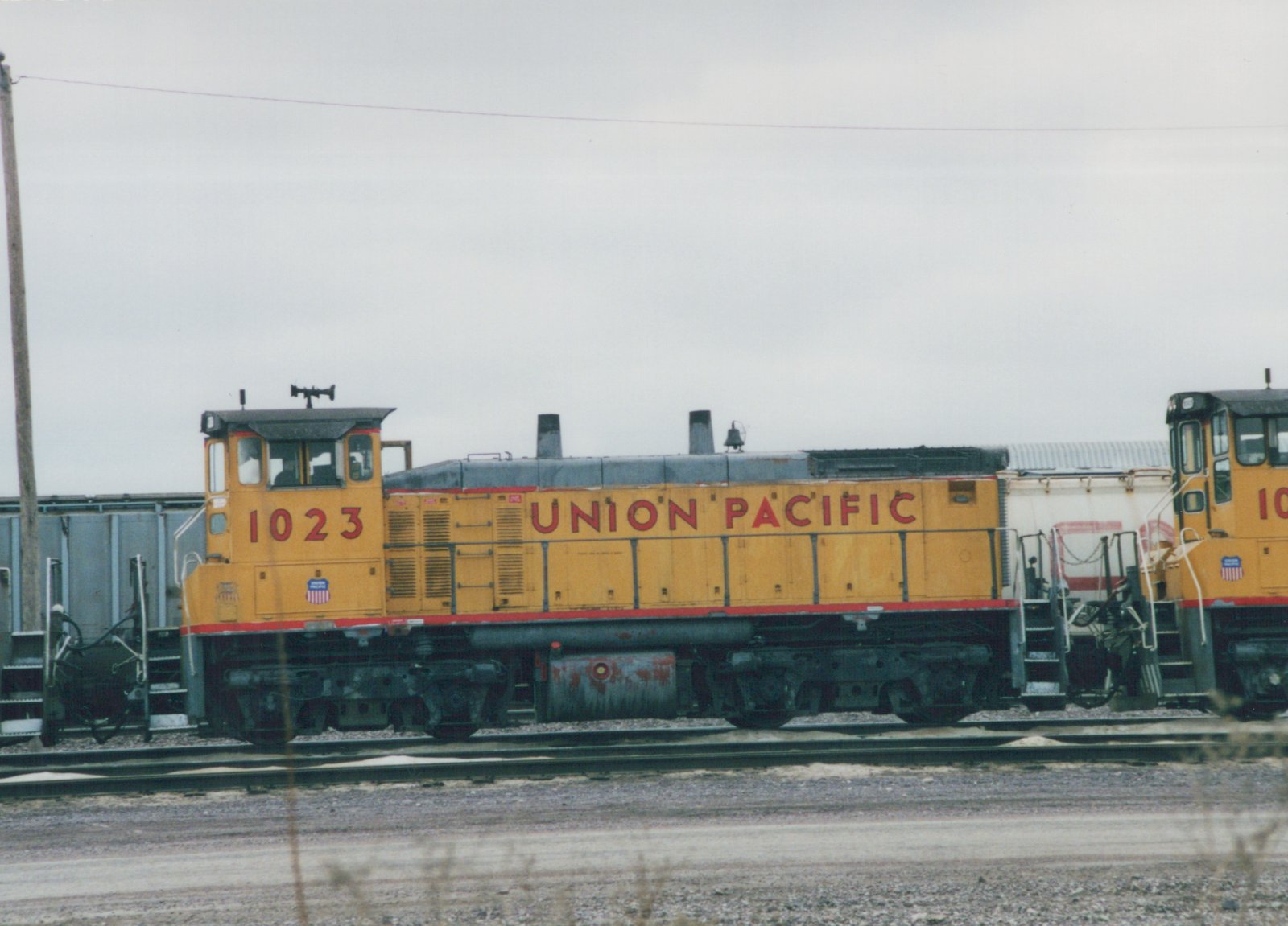 A yellow and gray Union Pacific diesel locomotive, numbered 1023, is parked on parallel tracks in a gravel yard under an overcast sky.