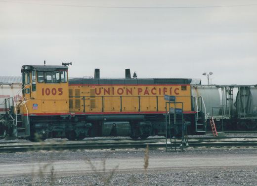 A side view of a yellow and black Union Pacific diesel locomotive numbered 1005, standing on tracks in a rail yard with freight cars behind it.