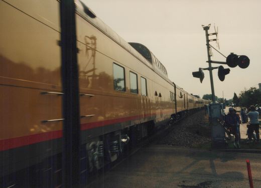 A long, tan and red bi-level passenger train, identified as an Amtrak Superliner, moves past a grade crossing with railroad signals, while several people on the sidewalk watch and take pictures.