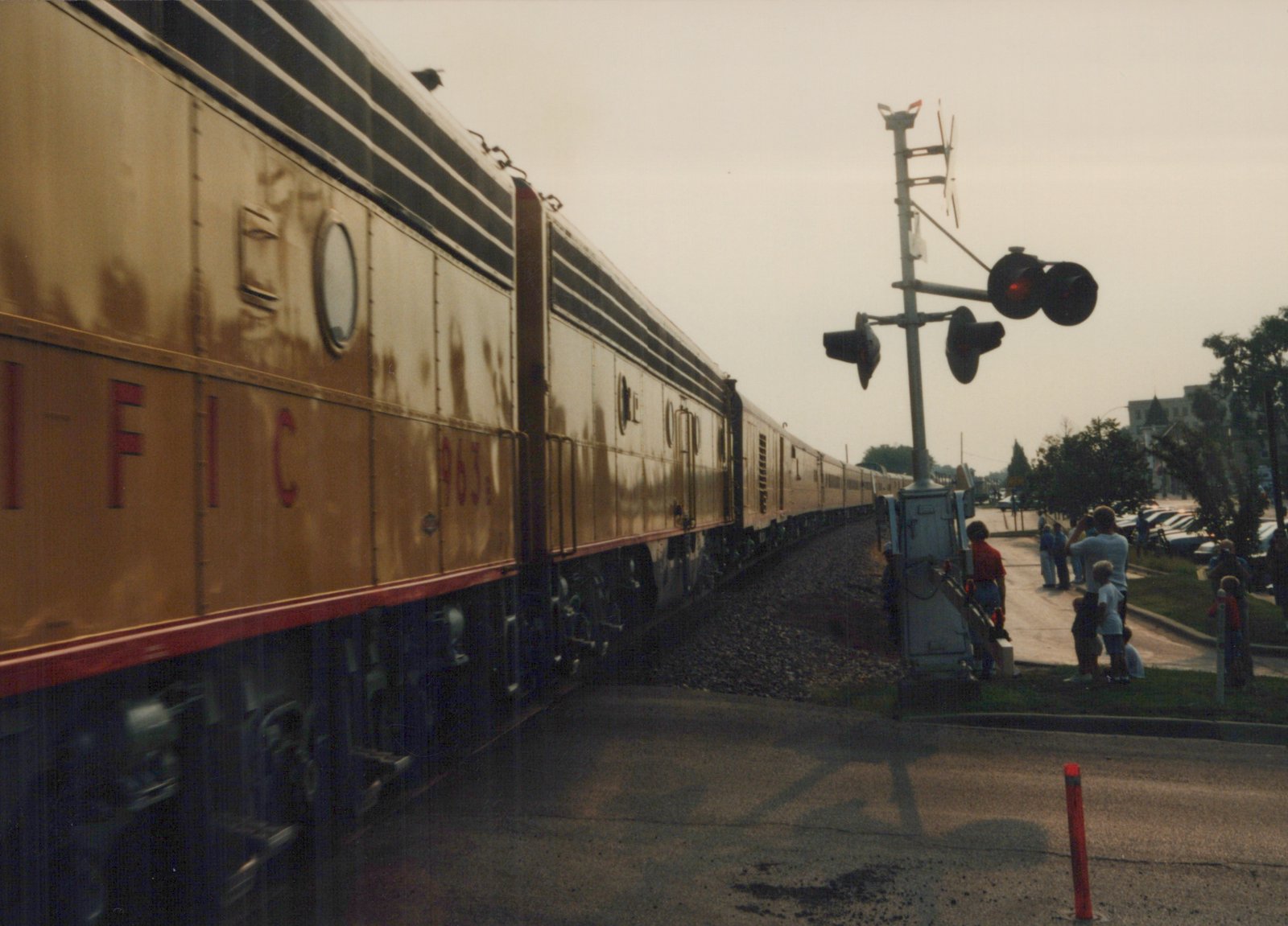 A gold and red CB&Q passenger train car numbered 963 moves past a grade crossing with several people watching from the side of the road.