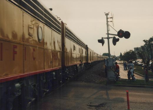 A gold and red CB&Q passenger train car numbered 963 moves past a grade crossing with several people watching from the side of the road.