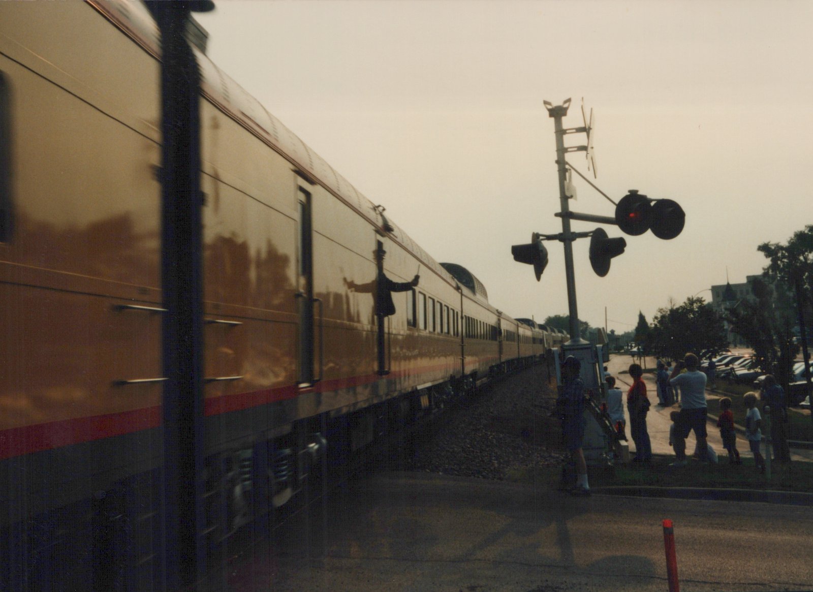 A long, two-level passenger train with a gold, red, and blue livery moves past a group of people standing near a railroad crossing signal in a suburban setting.