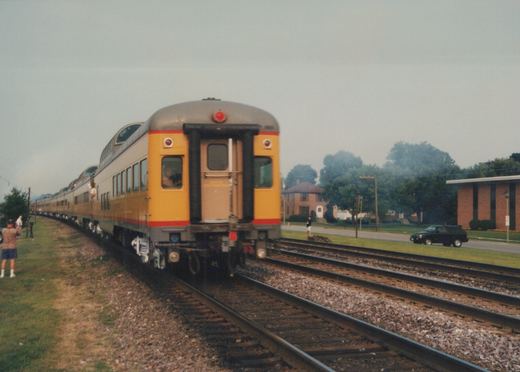 A yellow and red commuter car with double-decker windows travels on tracks next to a grassy area with people watching and a black SUV parked nearby.