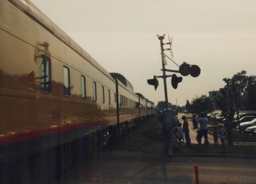 A long passenger train with a tan, dark blue, and red livery moves past a group of people standing at a grade crossing. The train features distinctive fluted sides and rounded roofline. Cars are parked in the background under an overcast sky.
