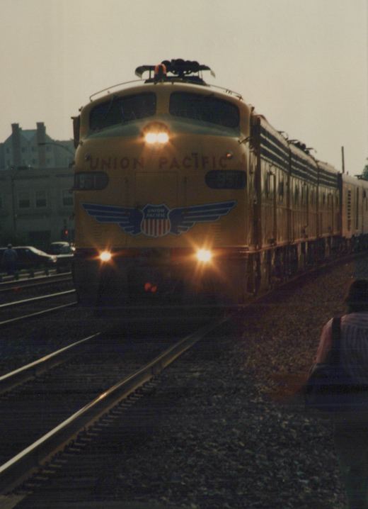 A yellow Union Pacific electric locomotive with number 951 on the front, pulling a passenger train, is seen approaching on railroad tracks at dusk, with a person in the foreground and buildings in the background.