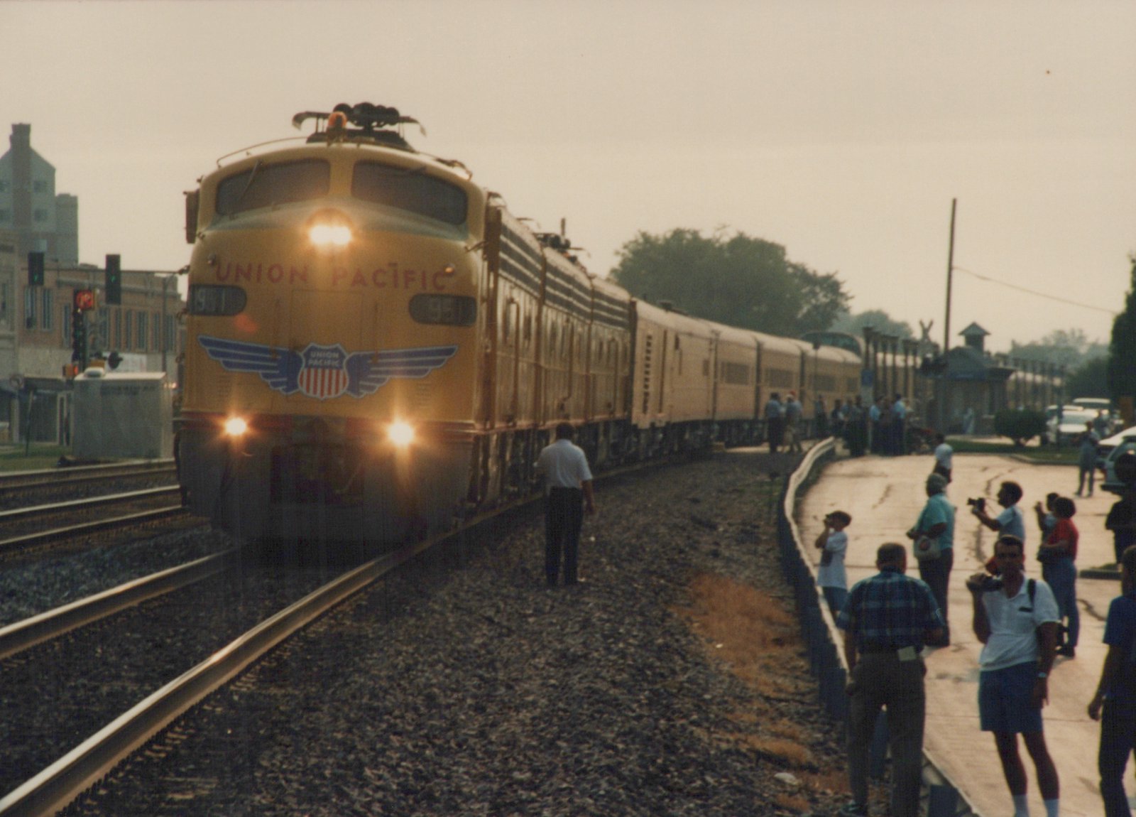 A yellow Union Pacific electric locomotive with its headlights on pulls a train past a crowd of people on a station platform on an overcast day.