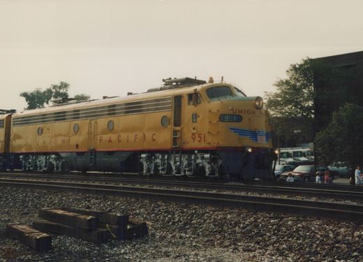A yellow Union Pacific E9 diesel locomotive numbered 951 is on railroad tracks with several people watching from the side and cars parked in the background under an overcast sky.