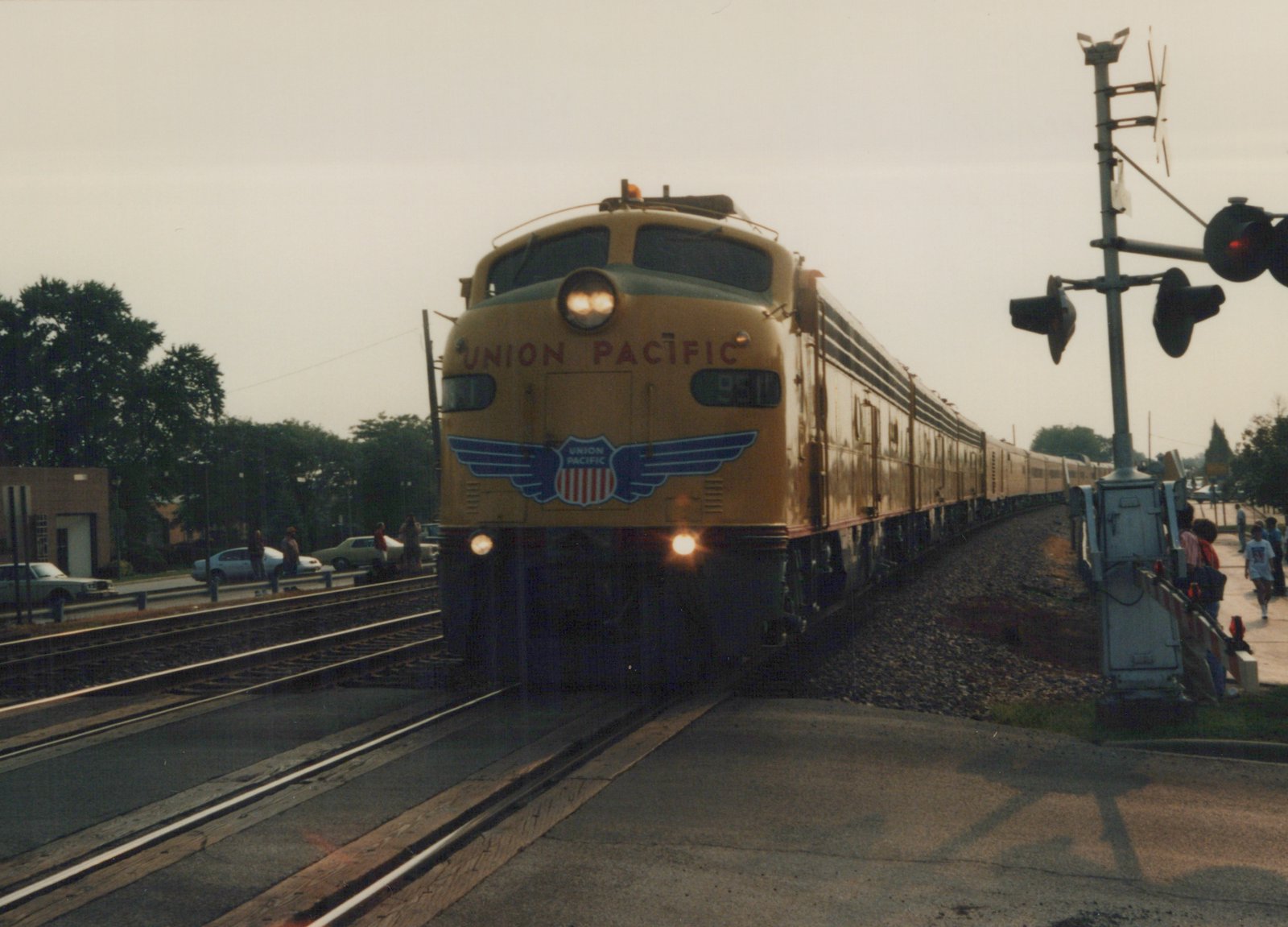 A yellow Union Pacific E9 locomotive numbered 951 pulls a long passenger train past a railroad crossing with people and cars nearby on a cloudy day.