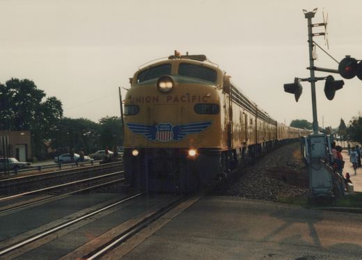 A yellow Union Pacific E9 locomotive numbered 951 pulls a long passenger train past a railroad crossing with people and cars nearby on a cloudy day.