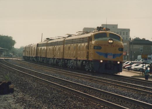 A yellow Union Pacific electric locomotive, number 951, moves along multiple railroad tracks with buildings and parked cars in the background, under an overcast sky.