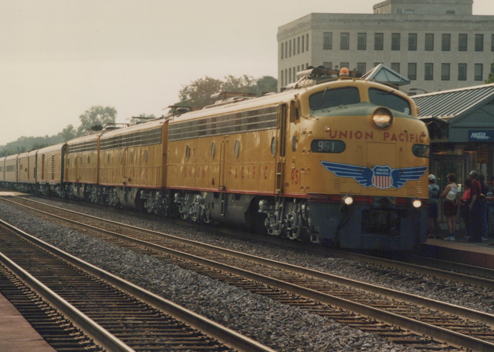 A yellow Union Pacific diesel locomotive numbered 951 is stopped at a station platform with a METRA sign. Several people wait on the platform as the train stands on the tracks.