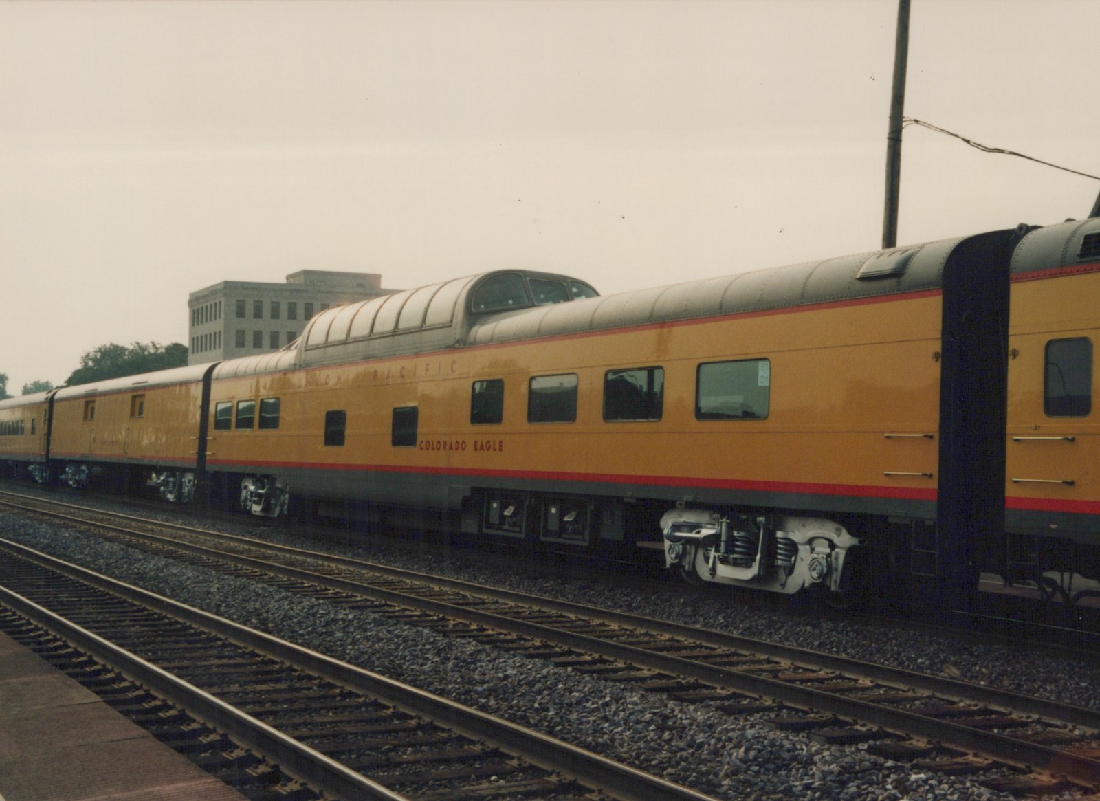 A side view of a Union Pacific Super Dome passenger car in yellow, grey, and red livery, sitting on railroad tracks next to a station platform under an overcast sky.