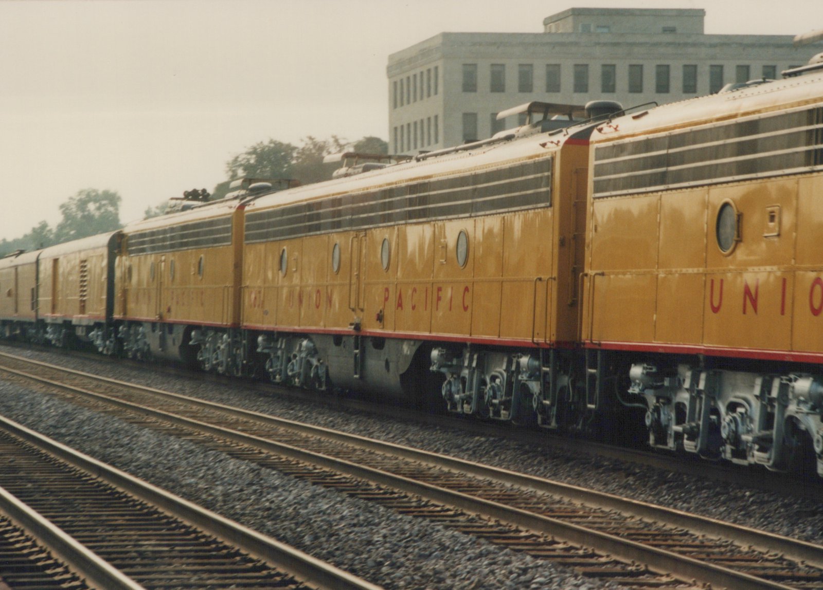 A yellow Union Pacific locomotive with electric pantographs travels on tracks beside a modern building, with passenger cars trailing behind.