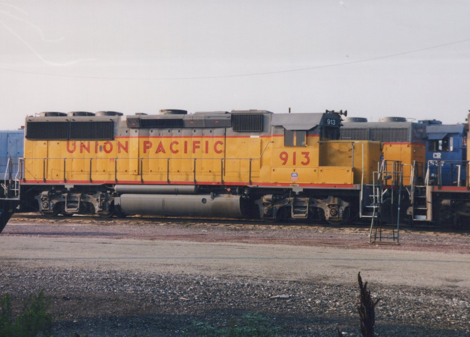 Side view of a yellow Union Pacific diesel locomotive numbered 913 in a rail yard, with a blue Chicago and North Western locomotive partially visible behind it.