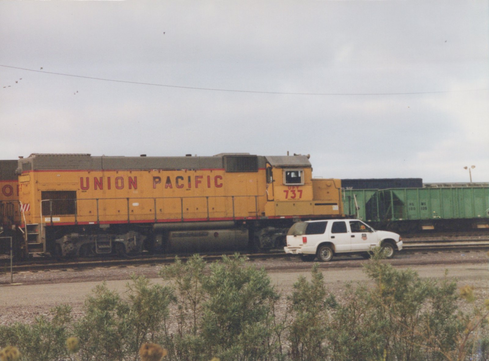 Side view of a yellow and gray Union Pacific diesel locomotive numbered 737 on a cloudy day, with a white SUV parked beside the tracks and green freight cars in the background.