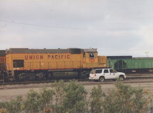 Side view of a yellow and gray Union Pacific diesel locomotive numbered 737 on a cloudy day, with a white SUV parked beside the tracks and green freight cars in the background.