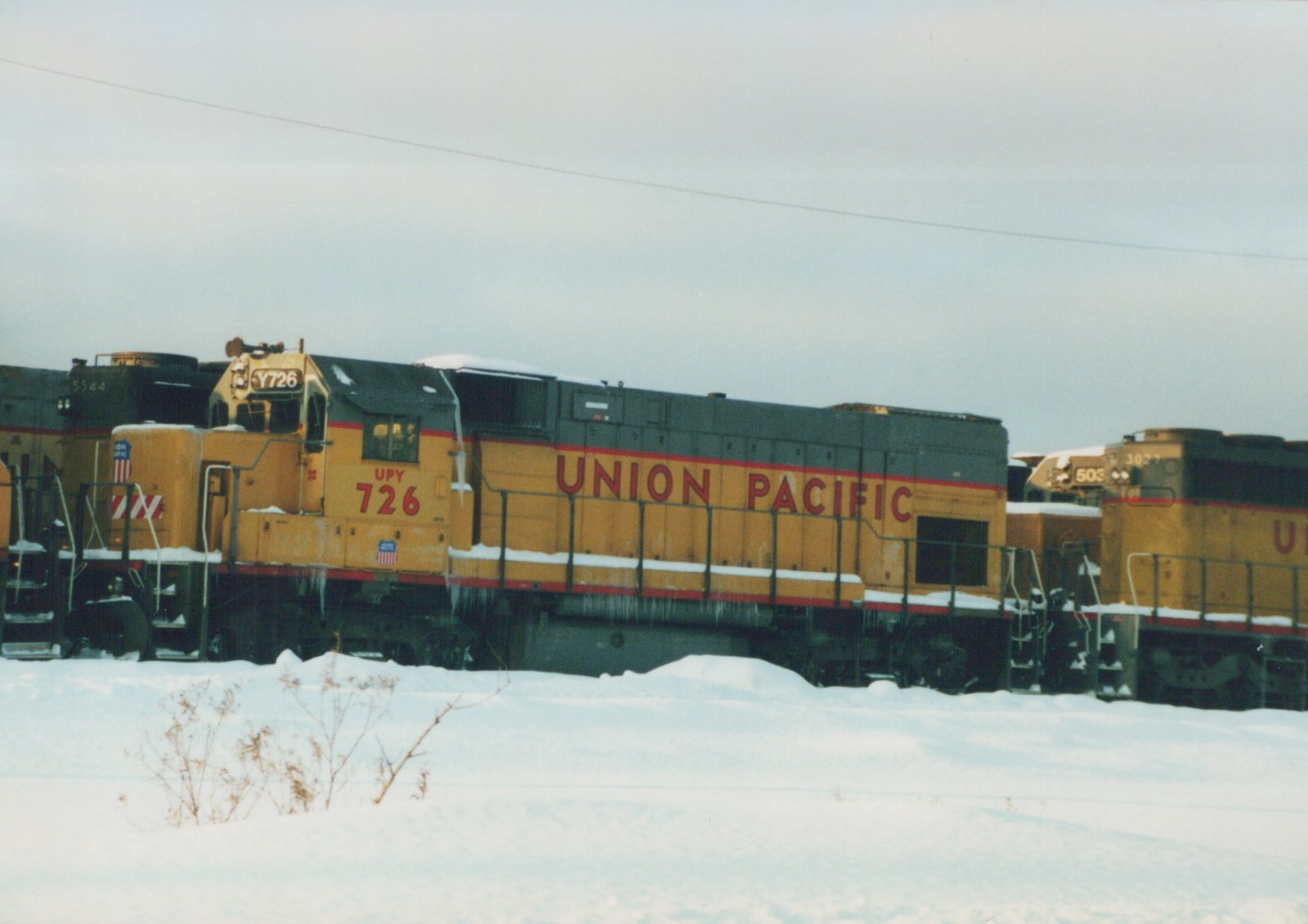 A yellow and gray Union Pacific diesel locomotive, number 726, stands in a snow-covered rail yard with other locomotives visible behind it.