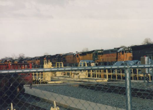A row of diesel locomotives, including Union Pacific 673 and Chicago and North Western 5505, are parked in a rail yard behind a chain-link fence on an overcast day.