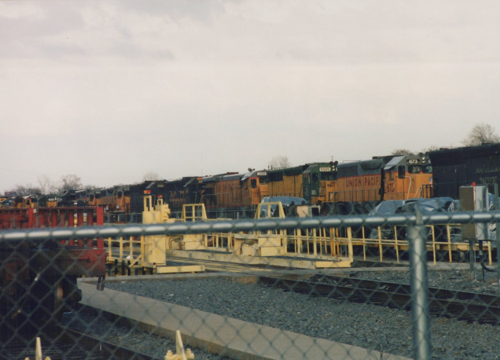 A row of diesel locomotives, including Union Pacific 673 and Chicago and North Western 5505, are parked in a rail yard behind a chain-link fence on an overcast day.