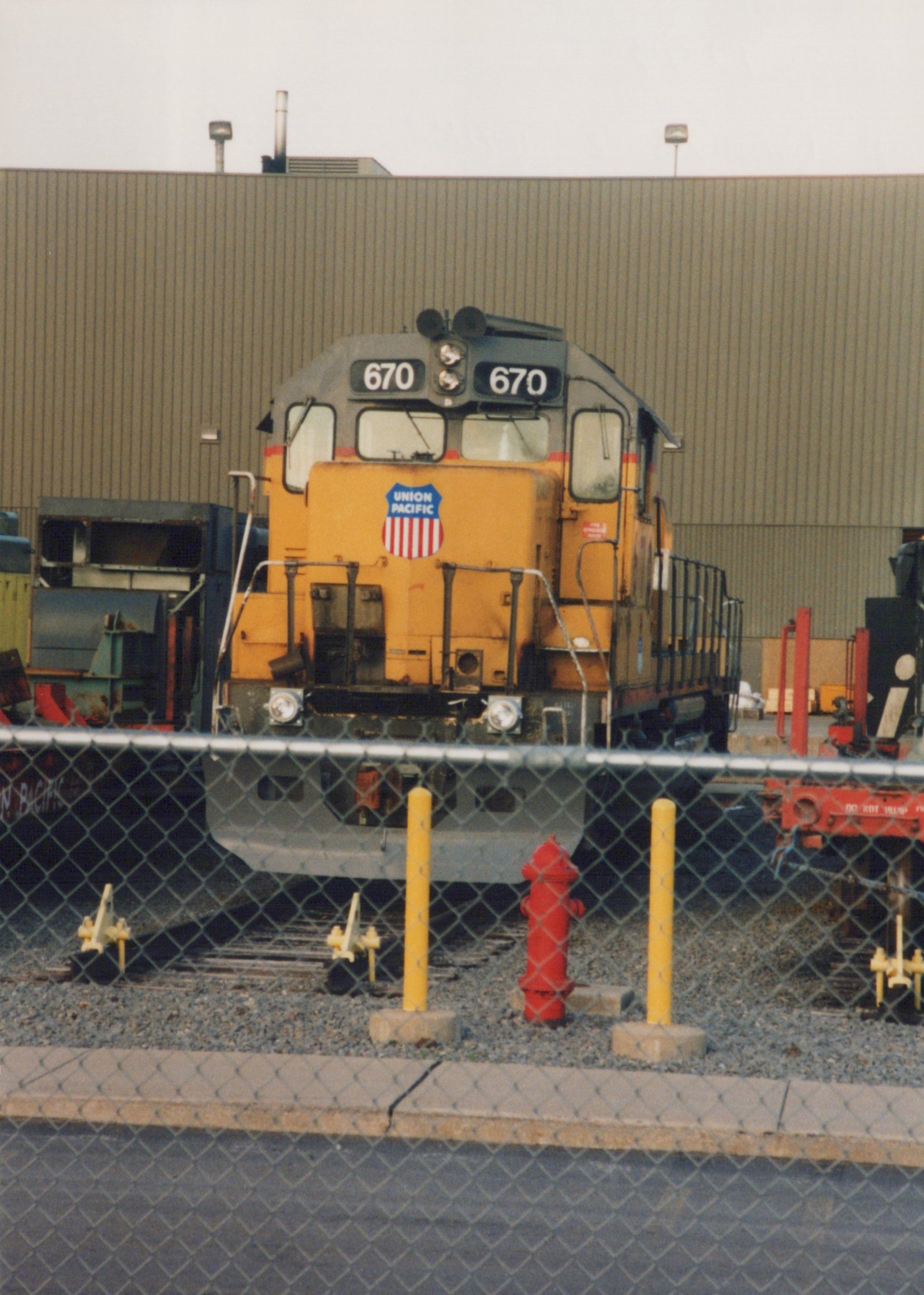 Front view of a yellow and grey Union Pacific diesel locomotive, number 670, parked on tracks behind a chain-link fence with a red fire hydrant and yellow bollards in the foreground.